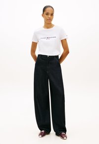 Frau steht mit den Händen hinter dem Rücken, trägt ein weißes Tommy Hilfiger T-Shirt, weite schwarze Jeans und weinrote Turnschuhe vor weißem Hintergrund.