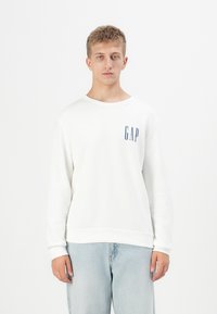 Sudadera blanca hecha de tejido suave, con mangas largas, cuello redondo y un logotipo azul de "GAP" en el lado izquierdo del pecho.