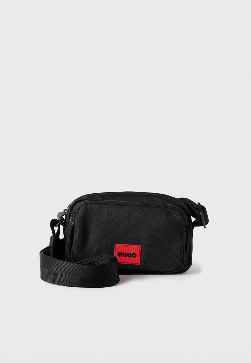 Black fabric crossbody bag with a textured surface, zip closure, and a rectangular shape. Features a red branded label and adjustable strap.