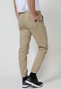 Bărbat purtând pantaloni jogger bej cu manșete elastice, tricou alb, șosete negre vizibile și adidași negri cu talpă albă, pe un fundal simplu deschis.