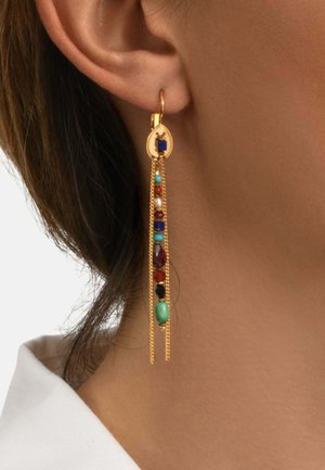 Boucle d'oreille goutte en or avec perles multicolores et chaînes en or délicates portée à l'oreille d'une femme aux cheveux bruns et col blanc visible.