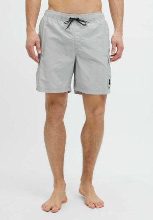 Homme portant un short gris clair, juste au-dessus du genou, avec un cordon noir, debout pieds nus sur un fond blanc.