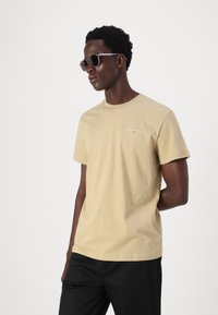 Beige cotton t-shirt with short sleeves, round neckline, and a small printed logo on the chest. Paired with black trousers and sunglasses.
