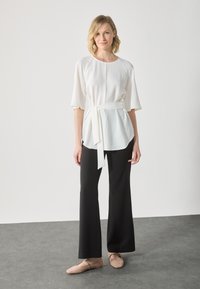 Top bianco, ampia e con maniche corte, con cintura annodata, abbinato a pantaloni neri a palazzo e scarpe basse color marrone chiaro su una superficie grigia.