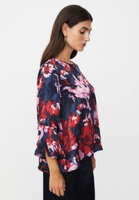 Bluse mit floralen Mustern, die einen dunkelblauen Grundton mit roten und pinken Motiven hat, dreiviertellange Ärmel mit gefransten Kanten und einen runden Halsausschnitt.
