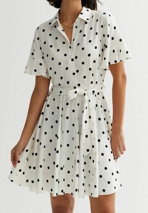 Robe blanche à manches courtes avec des pois noirs, un col, des boutons sur le devant et une ceinture nouée à la taille portée par une personne.