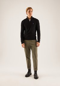 Pull en tricot noir avec col zippé, associé à un pantalon fuselé vert olive et des bottes noires à lacets, affichant une silhouette ajustée.