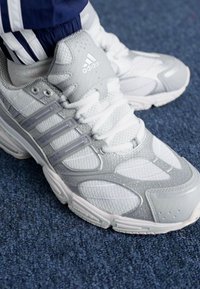 Adidas bėgimo batai šviesiai pilkos ir baltos spalvų. Tinkliniai ir sintetiniai medžiagos, tekstūruoti akcentai, ir trys skirtingos šoninės juostos ant viršaus.
