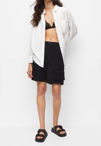 Femme portant une chemise blanche déboutonnée sur un soutien-gorge en dentelle noire, un short cargo noir et des sandales à boucle noires, se tenant contre un fond blanc.