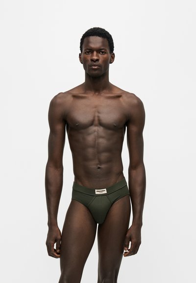 Homme musclé debout devant un fond uni portant un slip vert foncé avec une étiquette de marque sur la ceinture.
