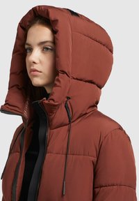 Červená puffer bunda s veľkou kapucňou, ktorá má prešívané časti, zips a šnúrky na nastavenie. Textúrovaná tkanina.