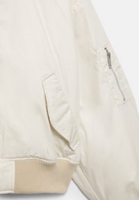 Jachetă bomber de culoare alb-crem, vedere apropiată care arată talia cu benzi elastice, buzunar cu clapetă pe corp și buzunar cu fermoar pe mânecă.