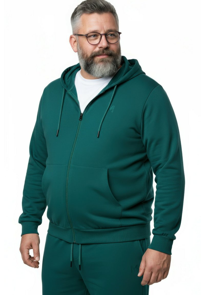 Uomo di mezza età con barba grigia che indossa occhiali, una felpa con zip color verde acqua, pantaloni abbinati e una maglietta bianca, che guarda leggermente di lato.