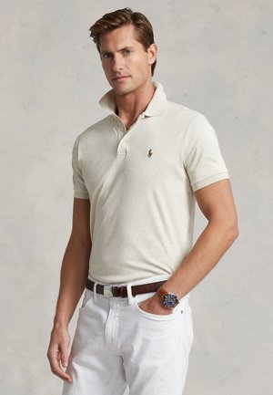 Mężczyzna ubrany w kremową koszulkę polo z logo, białe spodnie, brązowy pasek i brązowy zegarek na nadgarstku, stojący z jedną ręką w kieszeni na neutralnym tle.