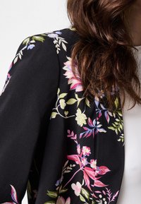 Close-up of a person wearing a black floral-patterned blazer over a white top, with long wavy brown hair partially visible.