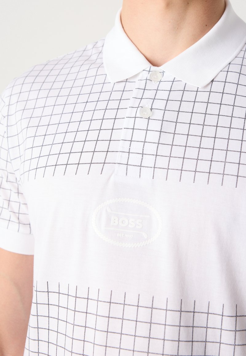 Camisa polo blanca con un patrón de cuadrícula en líneas negras y un logotipo en relieve en el pecho. Mangas cortas con un cuello clásico.
