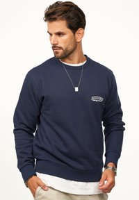 Navy pullover sweatshirt with ribbed texture, crew neckline, and a small logo on the chest. Layered over a white shirt.