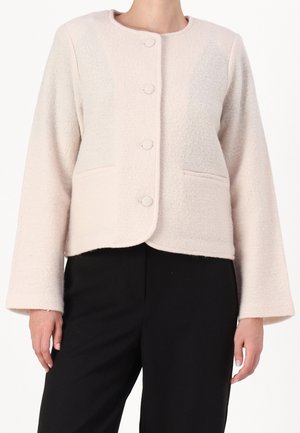 Vrouw draagt een lichtbeige textuur jasje met knopen, lange mouwen en zijzakken, gecombineerd met een zwarte broek.