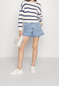 Jersey de punto a rayas en crema y azul marino, diseño corto con detalle de botones. Se lleva con shorts de denim azul claro que presentan costuras decorativas y bolsillos.