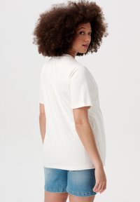 Hvid bomulds-T-shirt med korte �ærmer, afslappet pasform og rund halsudskæring, parret med lyseblå denimshorts. Modellen har krøllet hår.