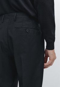 Zwarte op maat gemaakte broek met een gestructureerde afwerking, voorzien van een achterzak met een knoopsluiting, gedragen met een donkere trui.