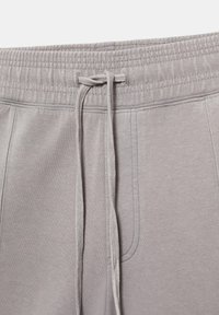 Pantalon de jogging gris clair avec taille élastique et cordon noué au centre devant, montrant des coutures détaillées et la texture du tissu.