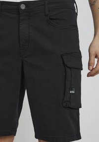 Shorts cargo noirs en tissu mélange de coton. Comprennent plusieurs poches, une fermeture à bouton et un écusson logo sur le côté.