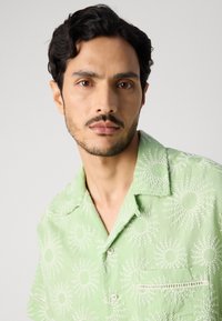Light green short-sleeve shirt featuring a sunburst pattern in white, with a breast pocket and subtle textured details.