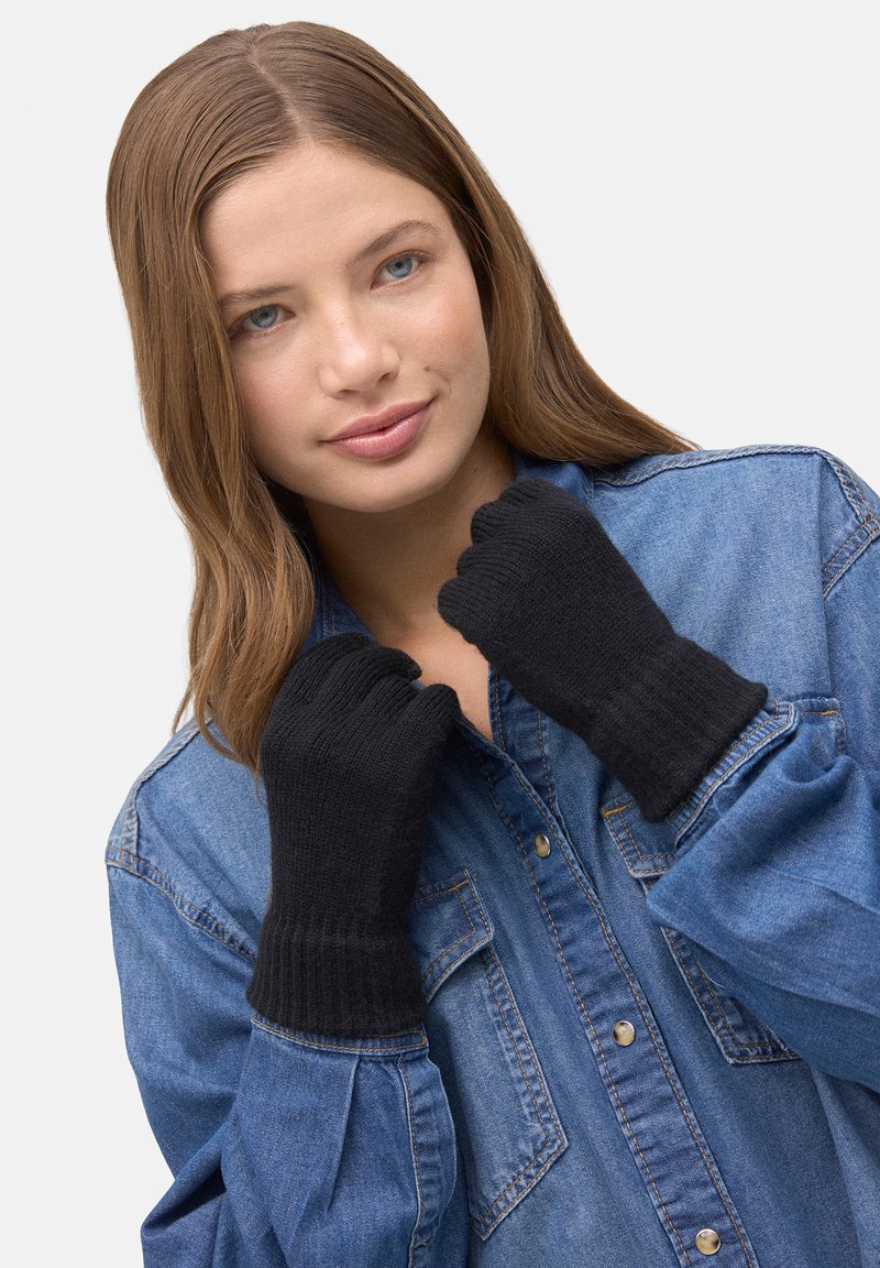 Guantes sin dedos de punto negro con puños acanalados, usados por una persona que lleva una camisa vaquera. Textura suave y diseño sencillo.