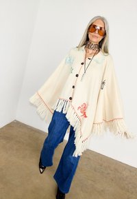 Γυναίκα που φοράει μπεζ πόαντσο με πολύχρωμη κέντημα, μπλε τζιν, καφέ φουλάρι και μεγάλα κίτρινα γυαλιά ηλίου, στέκεται σε τσιμεντένιο δάπεδο.