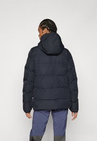 Mörk marinblå puffjacka med huva, med en quiltad design och synliga sömmar. Mjuk textur och överdimensionerad passform, lämplig för kallt väder.