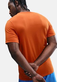 Camiseta naranja de manga corta, ajustada, en tejido suave, con un diseño simple de cuello redondo. La parte posterior presenta una textura lisa y sin gráficos visibles.