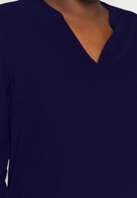 Top blu navy con scollatura a V e maniche lunghe, realizzato in tessuto morbido con una texture liscia, senza motivi o decorazioni visibili.