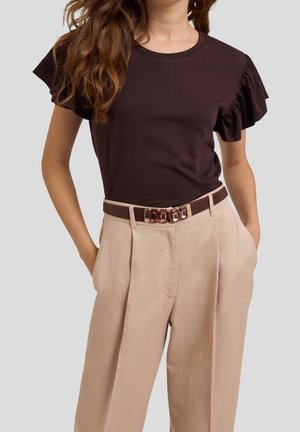 Vrouw draagt een donkerbruine top met ruches aan de mouwen, ingestopt in een beige broek met hoge taille en een bruine riem met een decoratieve gesp.