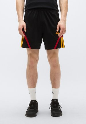 Pantaloncini sportivi neri con strisce laterali rosse e gialle, vita elasticizzata e una leggera lucentezza. Abbinati a scarpe da ginnastica nere e calzini bianchi.