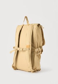 Μπεζ επενδυμένο σακίδιο με ρυθμιζόμενους ιμάντες, πλαϊνές τσέπες, πόρπη στο στήθος και κορυφαίο λουράκι για κρατησή, όρθιο σε απλό λευκό φόντο.