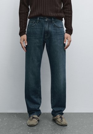 Dark blue denim jeans with a straight-leg design, featuring five pockets and subtle fading. Paired with light brown moccasins.