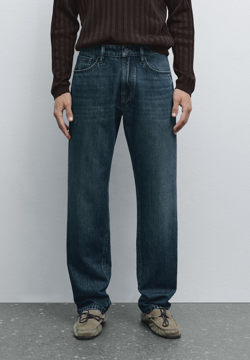 Jeans in denim blu scuro con design a gamba dritta, dotati di cinque tasche e un leggero sbiadimento. Abbinati a mocassini marrone chiaro.