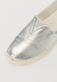 Silberne metallische Slipper mit runder Zehenpartie, die eine strukturierte Oberfläche und eine kontrastierende beige Sohle aufweisen. Dekorative Nähten sind sichtbar.