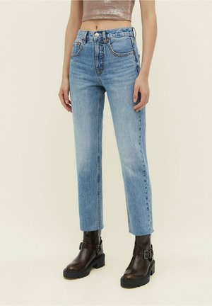 Femme portant un jean taille haute bleu clair à jambes droites, associé à des bottines noires à boucles, sur un fond uni.