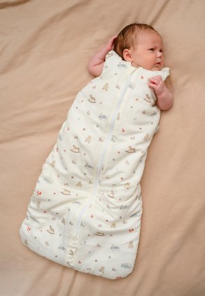 Vastasyntynyt makaa beigen värisellä viltillä, pukeutuneena valkoiseen unipussiin, jossa on pientä kuviointia teddykarhuista, keinuhevosista ja leluautoista.