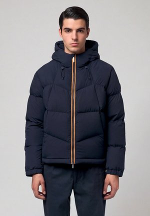 Giovane uomo che indossa una giacca a vento blu navy con cappuccio e cerniera arancione, in piedi contro uno sfondo chiaro uniforme.
