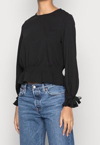 Schwarze kurz geschnittene Bluse mit langen Ärmeln, gerafftem Taillenbund und Rüschenakzenten an den Bündchen. Kombiniert mit hochgeschnittenen blauen Jeans.