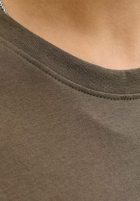 Close-up de la tela de una camiseta marrón de cuello redondo sobre la parte superior del pecho y el escote de una persona, mostrando detalles de textura y costura.