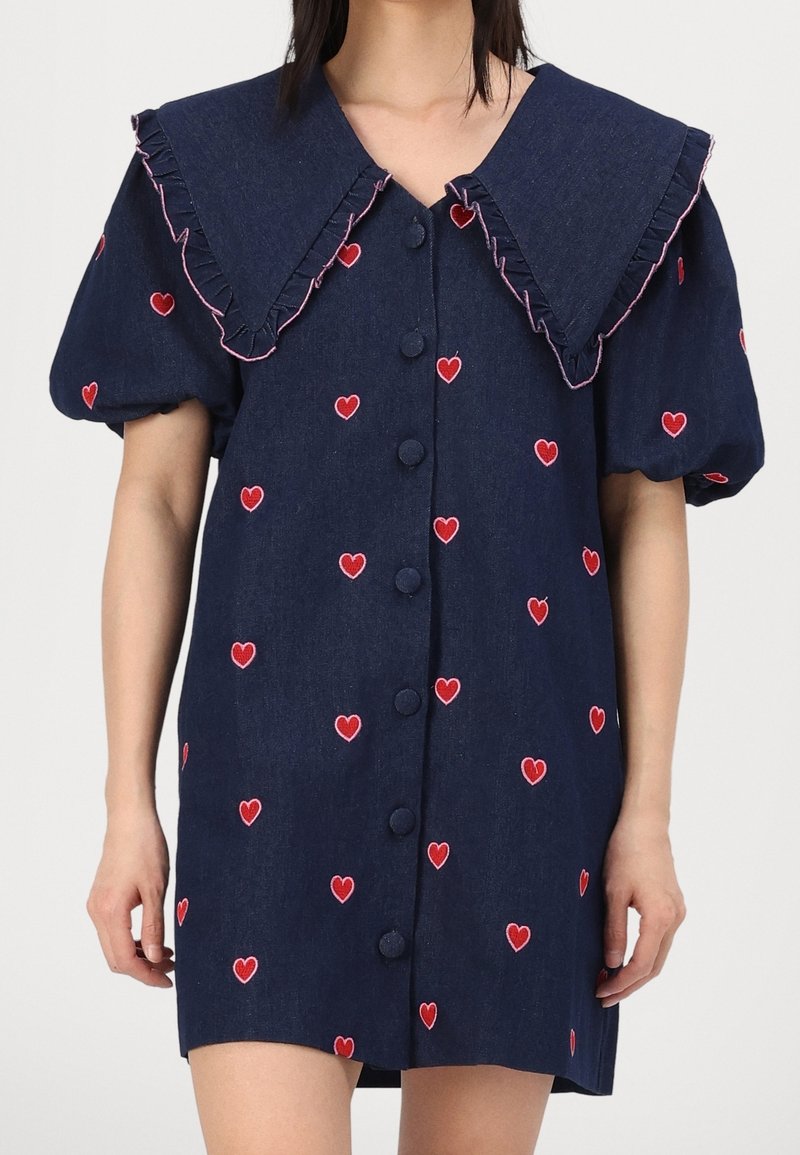 Femme portant une robe bleu marine avec des motifs de cœurs rouges, un grand col froncé avec un bord rose, des manches courtes bouffantes et des boutons recouverts de tissu sur le devant.