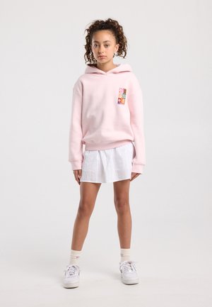 Meisje met krullend haar, draagend een lichtroze hoodie met kleurrijke patch, witte rok, crèmekleurige sokken en witte sneakers, staand tegen een witte achtergrond.