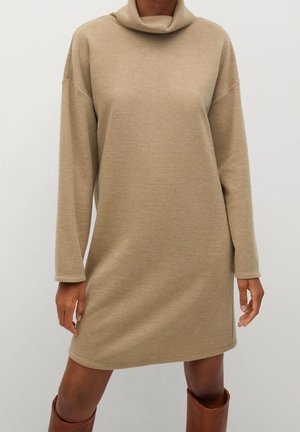 Beiges Mini-Kleid mit langen Ärmeln und lockerem Rollkragen, getragen mit braunen kniehohen Stiefeln, gezeigt an einem Model von Hals bis Mitte Oberschenkel.