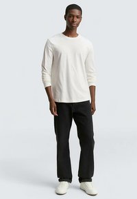 Kremowa koszulka z długim rękawem i okrągłym dekoltem, zestawiona z czarnymi spodniami o prostych nogawkach i białymi tenisówkami. Prosty design, miękka tekstura tkaniny.