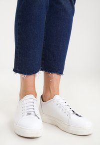 Zapatillas de cuero blancas con puntera redonda, suela plana y seis ojales. Combinadas con vaqueros de mezclilla azul oscuro con dobladillos deshilachados.