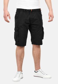 Black cargo shorts made of cotton with side pockets and a durable belt. Style features a straight cut and knee-length design. Worn with white sneakers.